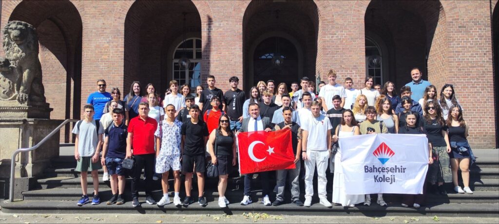 Gruppenfoto der Schülerinnen und Schüler der Bahcesehir Koleji Aspendos Kampüsü Antalya und Schülerinnen und Schülern der Erich-Fried-Gesamtschule Herne vor einem Gebäude. Die SchülerInnen halten eine türkische und eine Fahne mit dem Schulnamen in die Höhe.